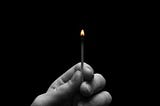 A black and white photo of a person holding up a lit match. The small fire is colored orange.