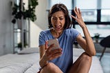 A middle-aged woman wearing a light-blue short sleeve t-shirt is sitting on her bed semi cross legged. She is looking at her phone and yelling, her phone is in her right hand. Her left hand is raised, with elbow resting on her left knee. Her facial expression reveals that she looks frustrated, confused, angry and maybe even bewildered.