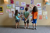 Three kids with their backs to the viewer, looking at dozens of drawings taped to the wall in front of them.