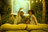 A Black couple take a selfie in their backyard, where they’ve set up a projector screen to watch a movie with hanging lights.