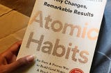 The book Atomic Habits by James Clear held in one hand over a slight grey background.