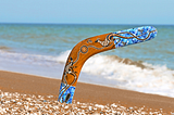 A colorful boomerang, one end of it stuck in the sand on a beach shoreline, symbolizing a rebound, personal growth, healing, reinvention, and life’s unexpected plot twists.
