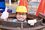 Workman with yellow hard hat and white gloves, rising out of a manhole giving the camera a thumbs-up.