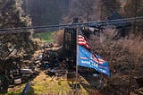 An American flag and a Trump flag flying over a burned down house.