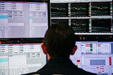 A photo that shows the back of a man’s head as he looks at stocks on four computer screens.