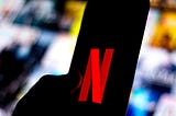 The Netflix logo is shown on a smartphone against a backdrop of blurred out Netflix show titles.