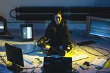 A woman in wearing a black jacket is sitting on the floor connected to various screens and keyboards.
