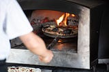 A person using a pizza peel to remove a pizza from an industrial metal wood-fired pizza oven.