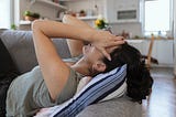 A person lying on their back on a couch in an open-concept living room/kitchen; their hands are over their face.