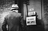 A black and white archival photo of a man standing in front of a door. The sign on the door says: ‘NO WORK. DON’T APPLY.’