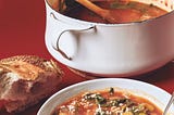 A pot of soup, a bowl of soup, and a small, rustic loaf of bread.