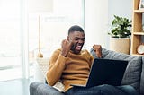 A young man cheers while using a laptop at home on his couch.