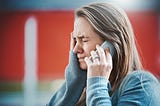 A young woman grimaces, her hand to her face as she hears something on the phone, obviously bad news.