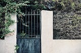 An iron gate in a stucco wall, covered in ivy and surrounded by trees and other overgrowth. The wall is parallel to a steep hill, but it is level, while the ground itself has a considerable slope.