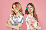 Two young ladies standing back to back wearing big silly sunglasses one looking put off and the other sticking her tongue out with an attitude