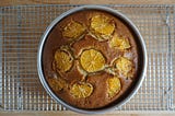 Overhead view of a cake still in its baking tin, resting on a wire rack. The cake is topped with 1 crosswise slice of orange in the center surrounded by 7 other slices.