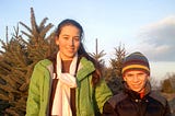 Two kids on a tree farm wearing winter wear; one holds a saw ready to take down a Christmas tree.
