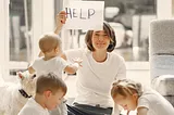 A disturbed mother holding a sign that says “Help” and surrounded by her three active children.