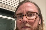 The author is wearing brown framed glasses and a pink shirt. He has a defiant look on his face. He’s standing in front of a window. It’s dark outside.