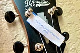 Closeup photo by the female story author of the top neck of a D’Angelico guitar, where a fortune cookie paper fortune strip lays on the strings that reads: “It is never a shame to learn from others.”