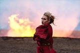 A woman in a red dress, standing in front of a fire looks back at the camera