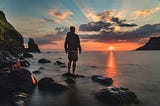 Standing on a small rock at the ocean’s edge, a lone man faces west and the orange glow of the sun at the horizen.