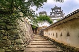 Stone pathway leading up to the main keep of Himeji Castle, flanked by traditional white plaster walls and stone defenses, with the iconic multi-tiered castle visible in the background.