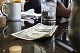 A close up of a dollar bill on a coffee table, next to a coffee mug and a glass of water.
