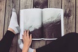 A photographer’s book is opened to a snowy mountain landscape as the reader’s legs and left hand are casually place around it while sitting on a wood patio deck.