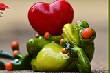 A green and yellow porcelain frog with orange bug eyes laying on its side with a big red heart balanced on his hip