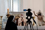 A nun with a video camera on a tripod recording a priest holding the Eucharist at an altar next to a statue of Jesus.