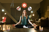 A woman meditates in lotus position on a yoga mat, surrounded by lit candles in a dimly lit living room. Above her floats an Om symbol and two theatrical masks. A red origami fox sits on a side table, with a voodoo doll in her left hand, and a meditation necklace in her right hand.