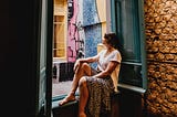 Barefoot woman sits on windowsill, gazing out open window onto a courtyard with street art