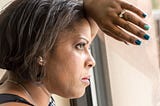 A closeup photo of a black woman leaning against a window and she looks out with a determined expression.