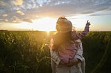 Mother holding daughter in a field at sunset with daughter pointing up to the sky