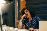 A young woman yawning at her desk while looking at her computer.