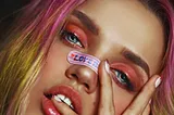 An airbrushed image of a woman with blue eyes and orange eyeshadow poses with her hand across her face. Her hair has pink roots and yellow ends, and a sticker over her nose reads “love”.