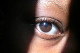 Closeup of a Black woman’s eye, with light shining in a strip where her eyes is. Rest of face obscured by shadow.