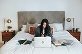 A person with a blanket draped around them sitting in bed working on a laptop, surrounded by books.