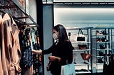 A person with long hair, wearing a mask and carrying one shopping bag, flips through clothes at a retail store.