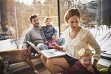 A photo of a family of four (mom, dad, two kids) sitting at home. Mom is on her computer and attending to a child.