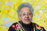 A portrait of Howardena Pindell in front of a colorful art piece.