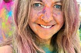 Midshot selfie of the female author smiling as she’s completely covered in the Holi Festival of Colors rainbow powders.