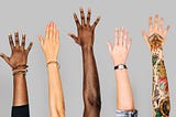 Six arms and hands raised up as if to volunteer, multiple ethnicities, set against a grey background.