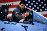Maverick is standing in the cockpit of his jet looking straight into the camera. He has his right hand in a thumbs up sign. He is wearing Naval Aviator attire, which is a dark grey; there are Naval patches on it. Written on the side of the jet is his call sign, Maverick. Behind him there is a blurred out American flag.