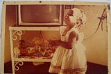 A young, Black girl of about two or three years of age looks up. Behind her is a television on a stand with a shelf below it on which a few dolls sit. The photo is from the 1980s.