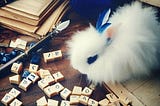 A fluffy white Blanc de Hotot rabbit sits contemplating blue and tan gaming tiles inscribed with strange letters, atop a wooden desktop also piled with old manuscripts and a quill pen.