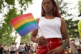 A young black woman holds up a rainbow flag at an outdoor gathering.