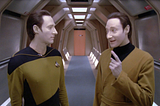 Brent Spiner as Lt Commander Data in a split screen with himself as Lore. Both are standing in a corridor on the USS Enterprise. Lore (on the right) is speaking, his face animated in a very human way. Data (on the left) listens, his face impassive and neutral.