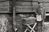 A sad-looking boy stands by a trestle table of newspapers, holding a copy for sale